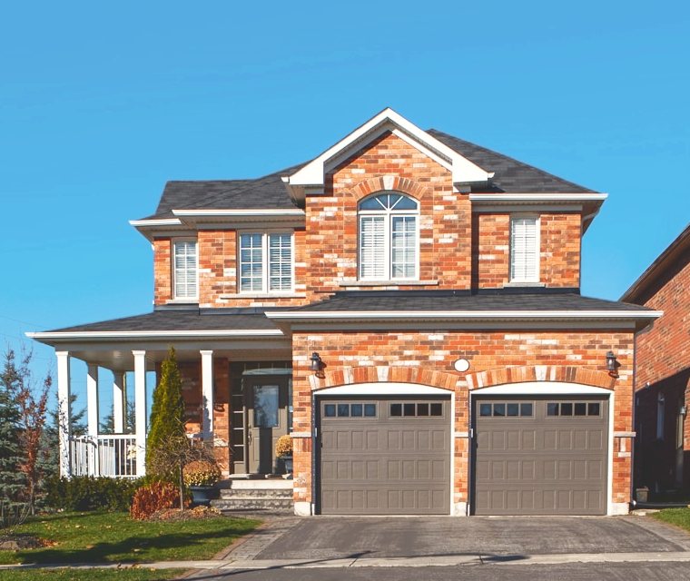 A two-story red-brick house with a gray shingle roof and a two-car garage under a clear blue sky